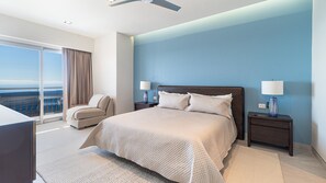 Ein modernes Hotelzimmer mit einem großen Bett, zwei Nachttischen mit Lampen, einem Sofa und Blick auf den Ozean durch eine Schiebetür.