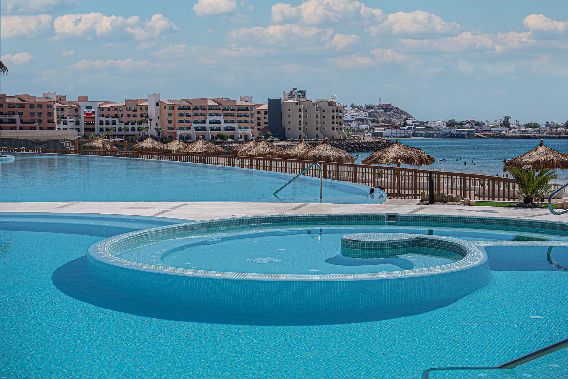 A large, clear swimming pool with a circular design, surrounded by a wooden deck and thatched umbrellas. In the background, there are multi-story buildings with balconies and a view of the sea.