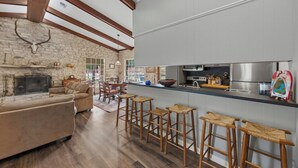 A spacious living area with a stone fireplace, a sofa, and a dining table with chairs. There are wooden stools at the kitchen counter. - Double H Ranch | Cozy 2-bedroom retreat + perfect for families! (Dripping Springs)
