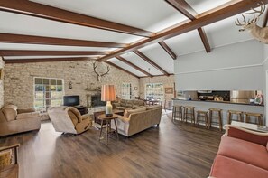 A spacious living room with a stone wall, wooden beams, a fireplace, and a kitchen area with stools. - Double H Ranch | Cozy 2-bedroom retreat + perfect for families! (Dripping Springs)