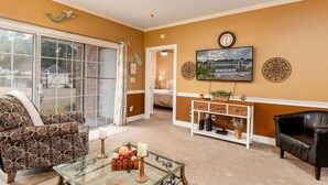 A living room with a glass coffee table, a sofa, a black armchair, a television mounted on the wall, and a ceiling fan. - Serene pool peaceful first floor kitchen washer dryer (Myrtle Beach)