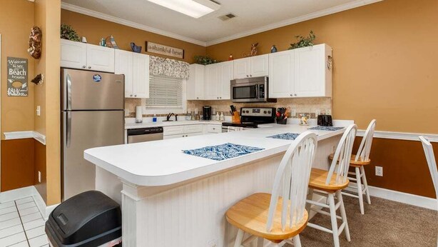 A kitchen with white cabinets, a stainless steel refrigerator, a microwave, and a central island with chairs. - Serene pool peaceful first floor kitchen washer dryer (Myrtle Beach)