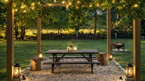 En træpergola med lyskæder, et picnicbord og lanterner i en have.