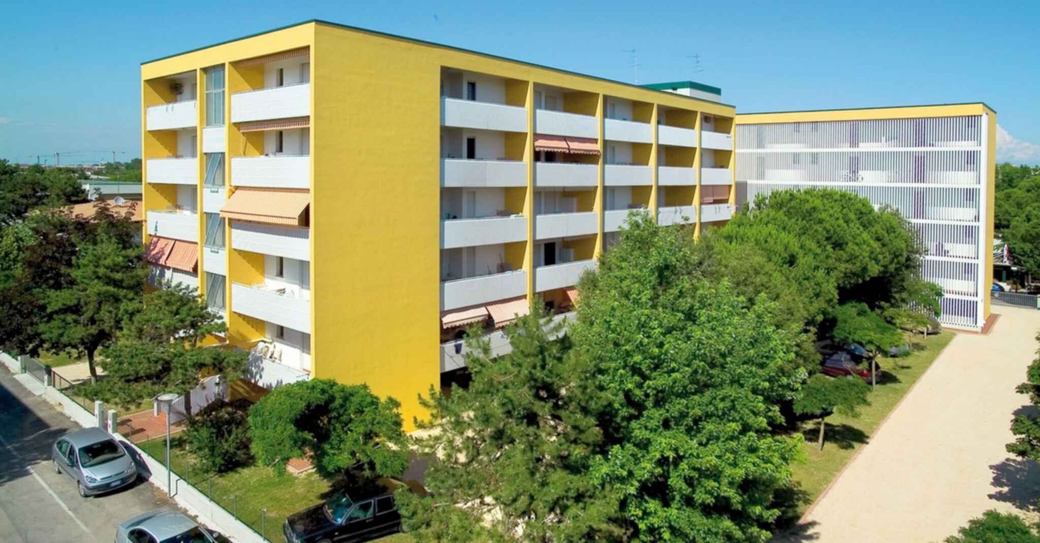 A multi-story building with a yellow facade and white balconies, surrounded by greenery and a clear sky.