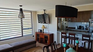 Ein Wohnzimmer mit einer Couch, einem Esstisch mit Stühlen, einem an der Wand montierten Fernseher und einem sichtbaren Küchenbereich durch eine offene Tür.