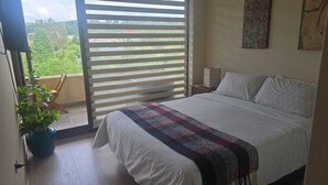 Ein Schlafzimmer mit einem Bett, einer Zimmerpflanze, einem Fenster mit Jalousien und Blick ins Grüne.