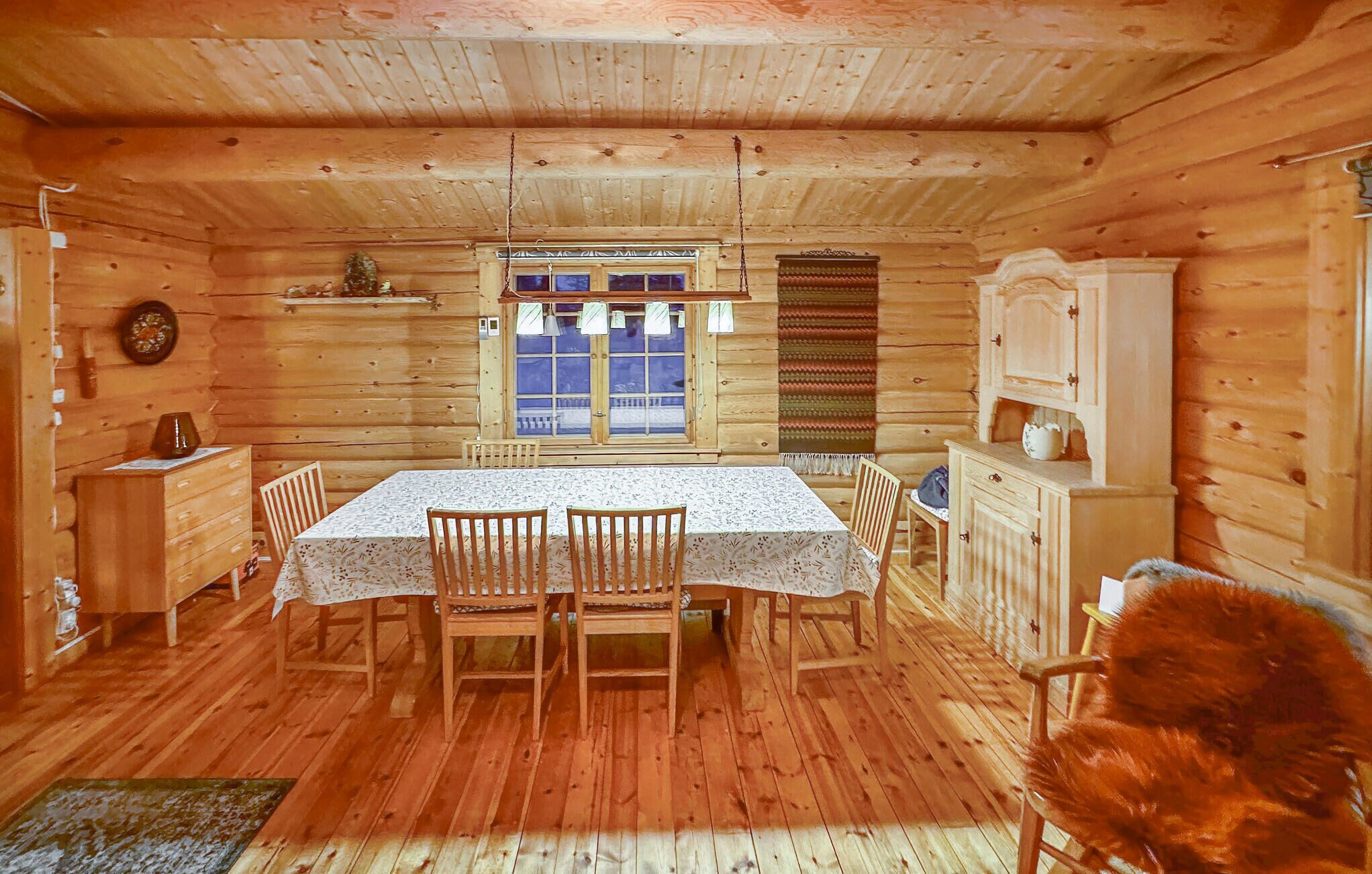 Een houten chalet van binnen, met een eettafel, stoelen, een kast en een raam met gordijnen.