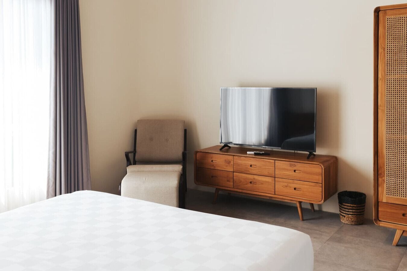 A hotel room with a bed, a wooden dresser with a television, a chair, and a wicker side table.