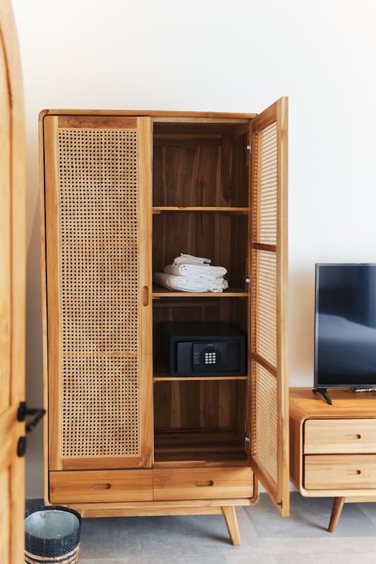 A wooden wardrobe with a woven front panel, a television on a wooden stand, and a small trash bin.