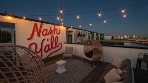 A rooftop terrace with a large wall featuring the text "Nashville," string lights, wicker chairs, a sofa, and a patterned rug. - The Bluebell - Music City Views, Game Room, 11 Beds! (Nashville)