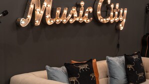 A room with a dark wall, a light-colored sofa with patterned cushions, and a neon sign reading "Music City". - The Bluebell - Music City Views, Game Room, 11 Beds! (Nashville)