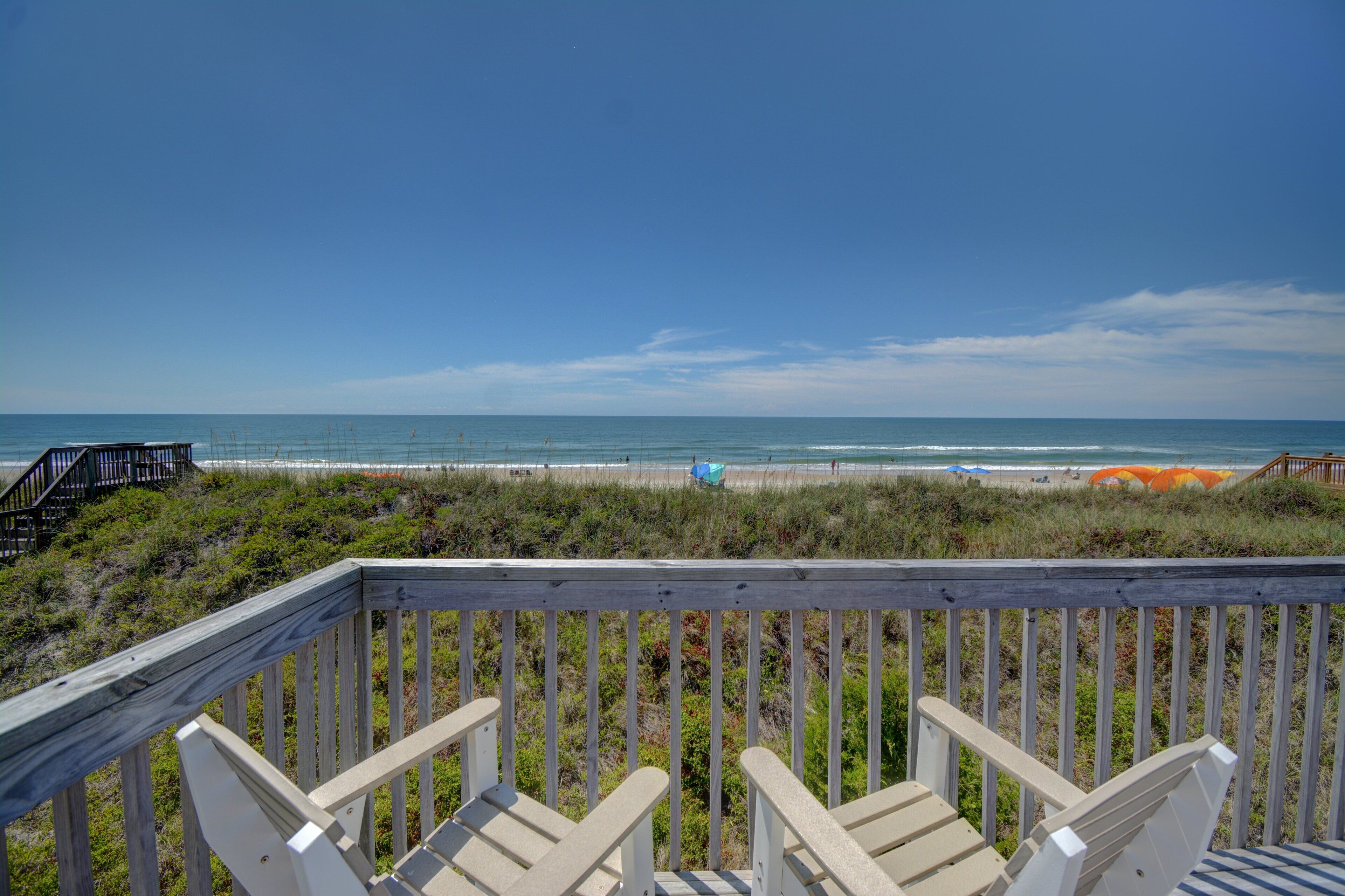 A wooden deck with chairs overlooking a beach with umbrellas and a staircase leading to the beach.