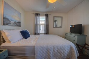 Une chambre à coucher avec un lit, un téléviseur posé sur une commode, un tableau accroché au mur et une fenêtre avec des rideaux.