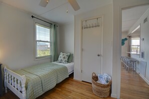 Ein Einzelbett mit grüner Steppdecke, ein Fenster mit Jalousien, eine Tür mit Kleiderhaken und ein Korbwäschetablett auf dem Boden.