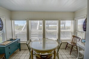 A dining area with a table and chairs, large windows with blinds, a blue dresser, and a chair with a decorative backrest. - Cozy 2BR retreat in North Topsail Beach, perfect for relaxing getaways (North Topsail Beach)