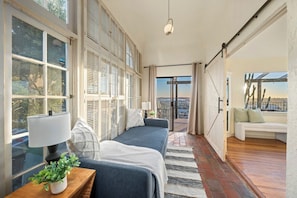 A room with a bed, a bedside table with a lamp, a window with blinds, a view through a glass door, and a sofa with cushions. - Prestigious Hills Estate | Stunning Pool & Hot Tub Views (San Diego)