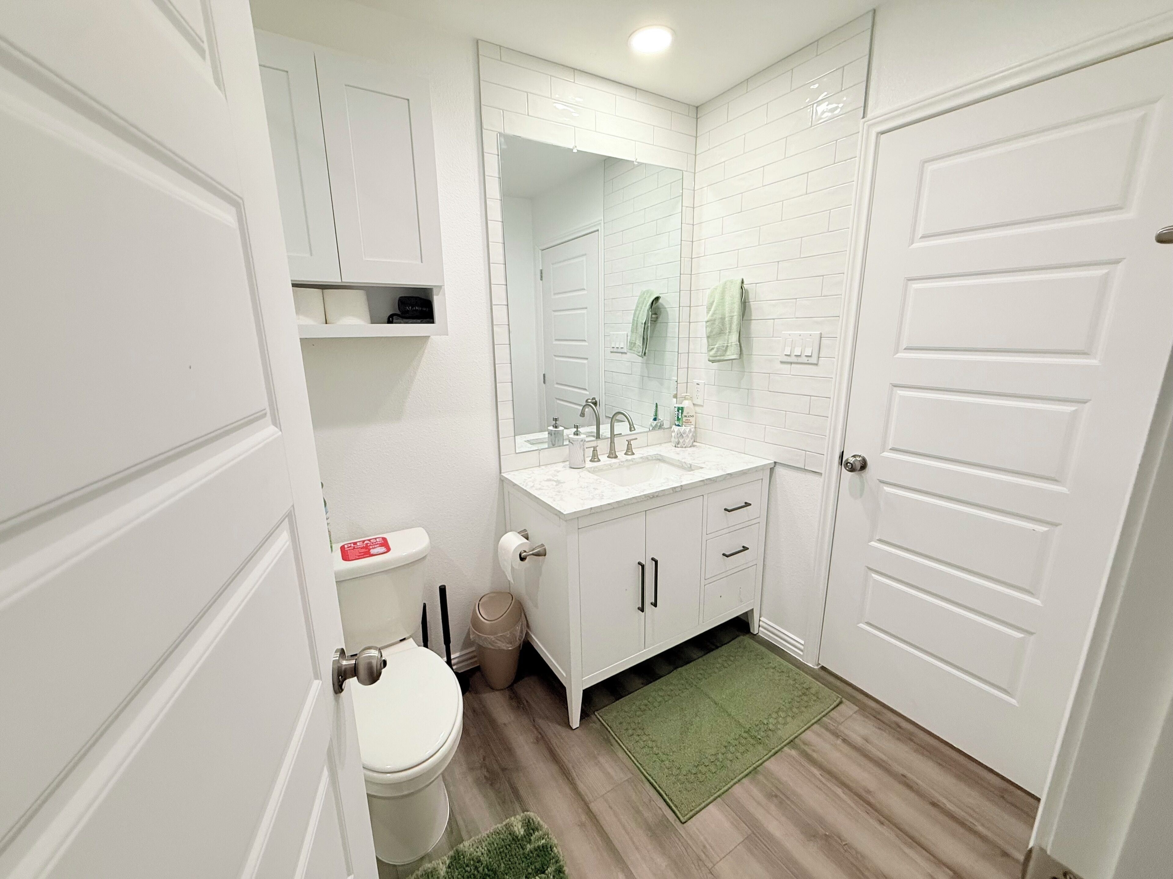 Un baño con muebles blancos, un inodoro, un lavabo con espejo y una alfombra verde.