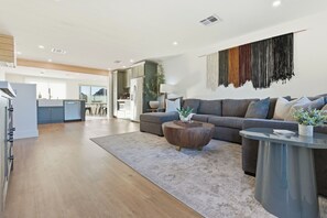 Ein modernes Wohnzimmer mit einem grauen Sofa, einem Holztisch und einem runden Glastisch. Im Hintergrund ist eine Küche mit einem grauen Schrank und einer weißen Arbeitsplatte zu sehen.