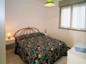 Ein Schlafzimmer mit einem Bett, einem Nachttisch, einer Lampe, einem Fenster mit Vorhängen und einem Schreibtisch mit Stuhl.