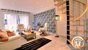Een moderne woonkamer met een bank, fauteuils, een salontafel en een televisie. Er is behang met een patroon en een trap met een houten leuning.