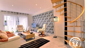 Ein modernes Wohnzimmer mit Sofa, Sesseln, Couchtisch und Fernseher. An den Wänden ist eine strukturierte Tapete und eine Treppe mit Holzgeländer.