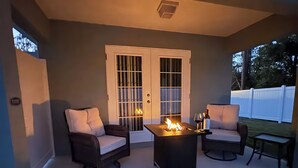 Eine überdachte Terrasse mit zwei Stühlen, einem Tisch mit Feuerstelle und Blick auf einen weißen Zaun.