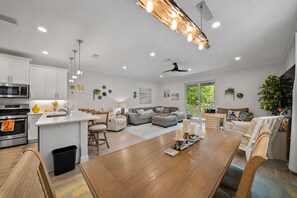 A modern kitchen with a central island, a dining area with a wooden table, and a living room with a sofa and a ceiling fan. - Modern Dunedin Home - Walk to Downtown, Near Beach (Dunedin)
