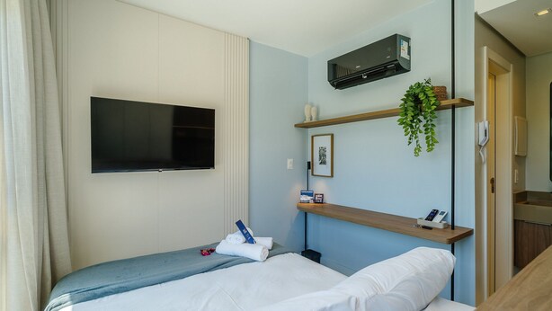 Ein modernes Hotelzimmer mit einem Bett, einem Wand-Fernseher, einem kleinen Regal mit einer Pflanze und einem Telefon.