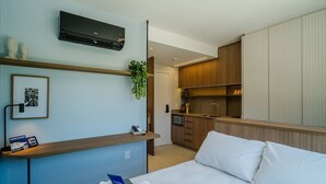 Ein modernes Hotelzimmer mit Bett, Nachttisch, Wandairconditioner, einer kleinen Küchenzeile und einem gerahmten Bild an der Wand.