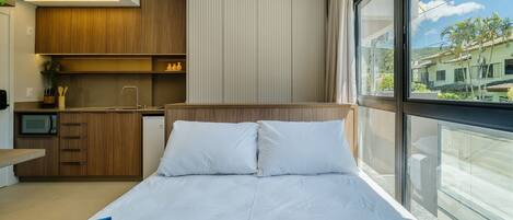 A modern hotel room with a bed, a small kitchenette, and a large window offering a view of the outside.