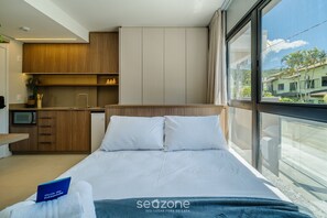 A modern hotel room with a bed, a small kitchenette, and a large window offering a view of the outside. - Apt Center Lagoa w/ Rooftop Pool Lst0002 (Florianópolis)