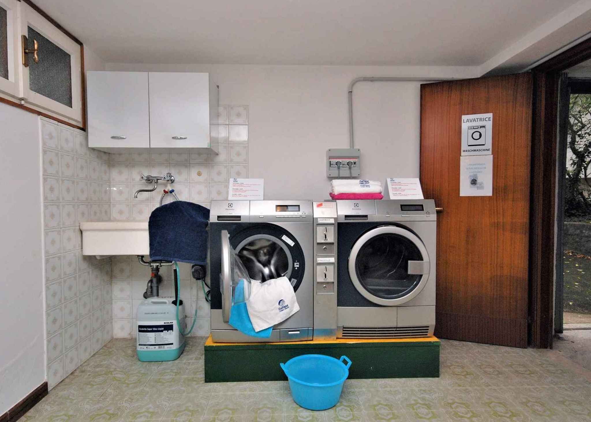 Lavandería con lavadora, secadora y lavabo. Hay un cubo azul en el suelo y un cartel en la pared.