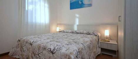 Un dormitorio con cama, dos mesitas de noche con lámparas, un cuadro en la pared y una ventana con cortinas.