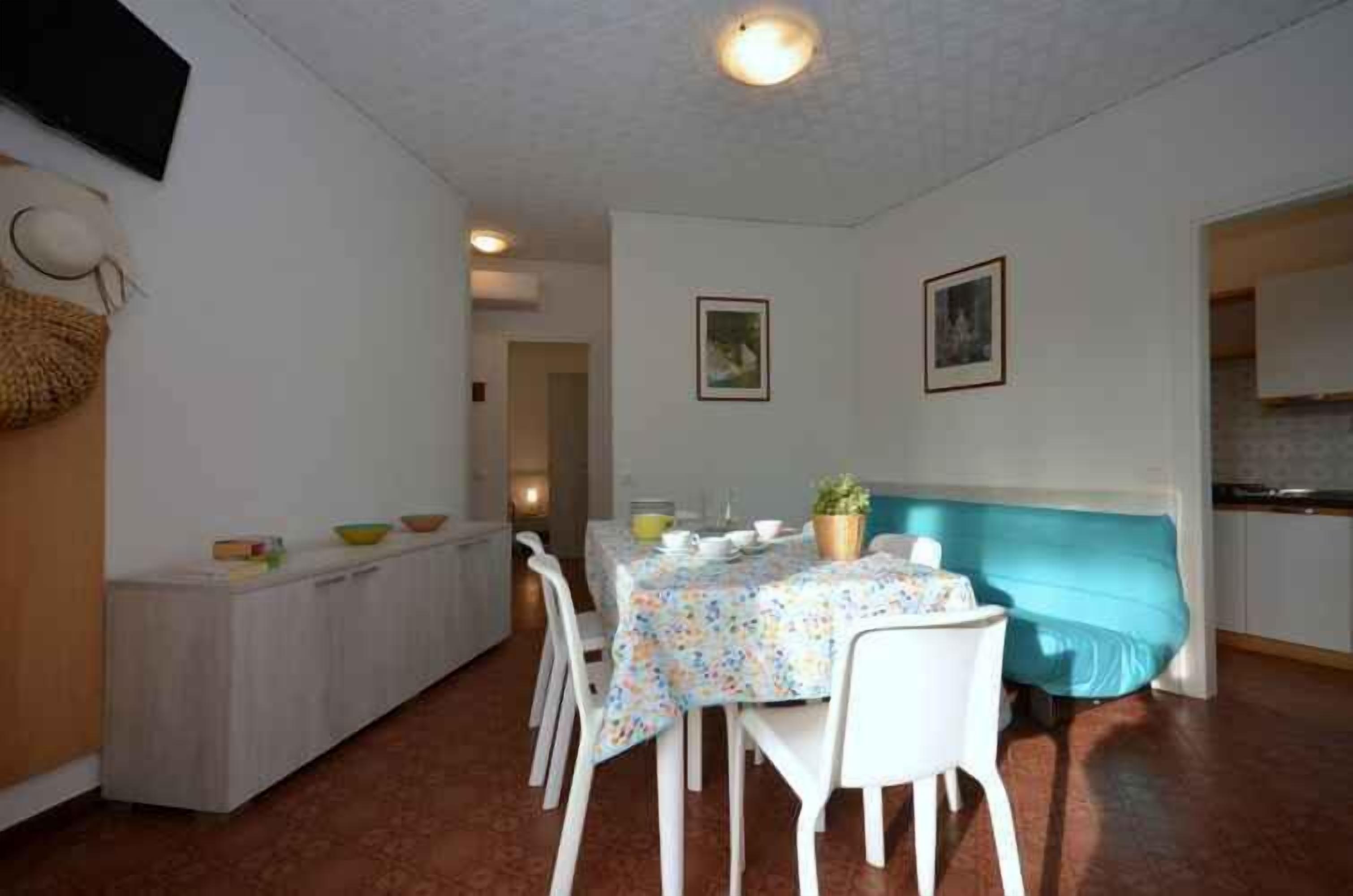 A dining area with a table set for four, white chairs, a blue-covered bench, and a kitchen in the background.