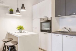 Une cuisine moderne avec des meubles blancs, un four encastré, un évier, des luminaires suspendus et deux tabourets.