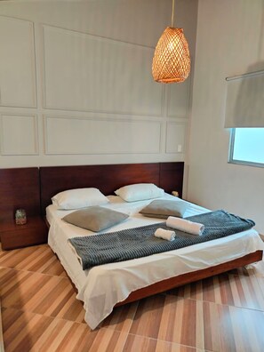 Ein modernes Schlafzimmer mit einem Holz-Kopfteil, einem großen Bett mit weißer Bettwäsche, einer gewebten Pendelleuchte und einem Fenster mit Jalousien.