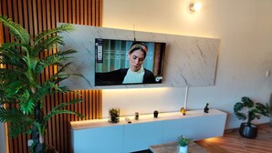 En moderne stue med et TV ophængt på væggen, et hvidt konsolbord, et lille sofabord og en stor potteplante.