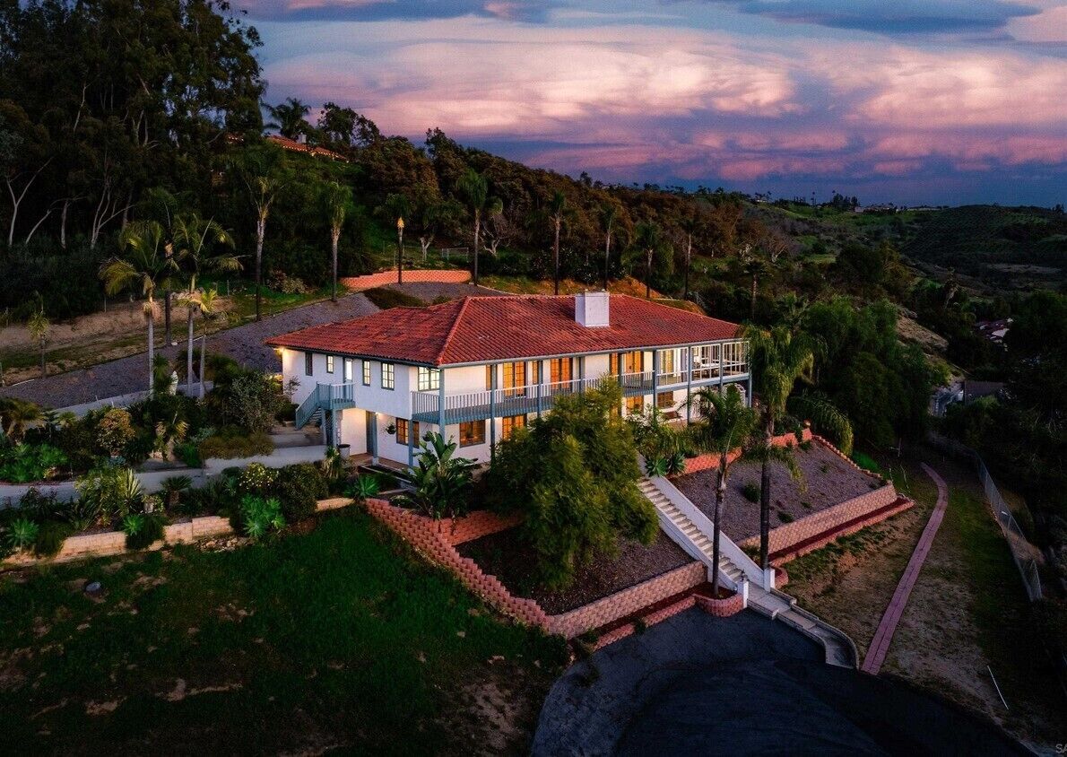 A two-story house with a red-tiled roof, surrounded by lush greenery and a hilly landscape.