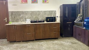 A kitchen with wooden cabinets, a sink, a microwave, and a refrigerator. There are three framed motivational posters on the wall. - Luxury 2BHK Villa with Private Pool – Sector 135, Noida (Noida)