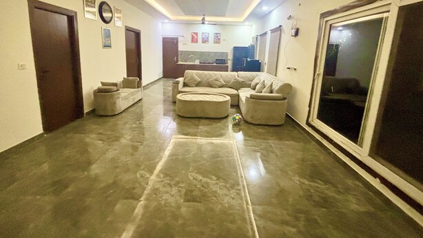 A spacious living area with a large sectional sofa, ottomans, and a dining area with a kitchen in the background. - Luxury 2BHK Villa with Private Pool – Sector 135, Noida (Noida)