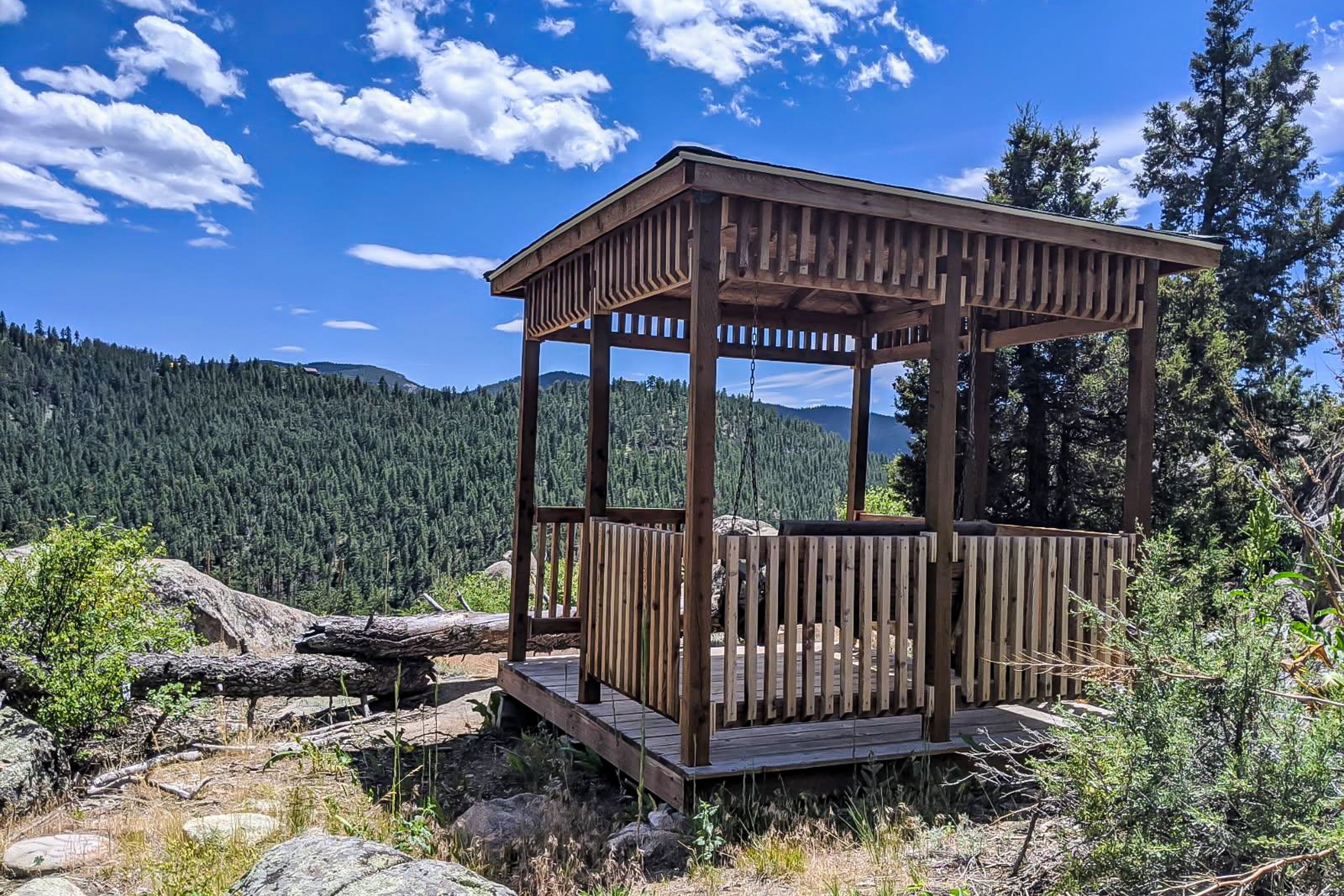 A wooden gazebo with a platform, surrounded by natural landscape and trees.