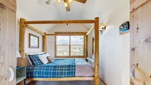 Una cama rústica con dosel de madera, con edredón de cuadros azules y blancos, una mesita de noche de madera con una lámpara y vistas al desierto a través de un ventanal grande.