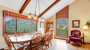 A dining area with a wooden table and chairs, large windows with a view of greenery, and a red armchair with a lamp. - 3BR with Epic Mountain Views and Huge Deck! (Estes Park)