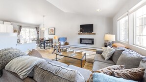 Ein geräumiges Wohnzimmer mit einer großen Couch, einem Couchtisch und einem Kamin. Ein Fernseher ist an der Wand befestigt und es gibt einen Essbereich mit Tisch und Stühlen.