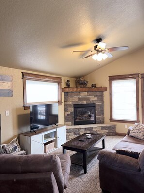 A living room with a stone fireplace, a ceiling fan, a flat-screen TV, and a sofa. - Cozy mountain townhome with deck, jetted tub, and gas fireplace (Estes Park)