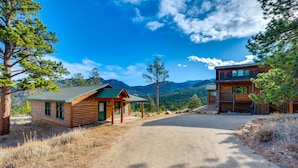 A rustic cabin with a gravel driveway, surrounded by pine trees and mountains.