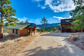 A rustic cabin with a gravel driveway, surrounded by pine trees and mountains.