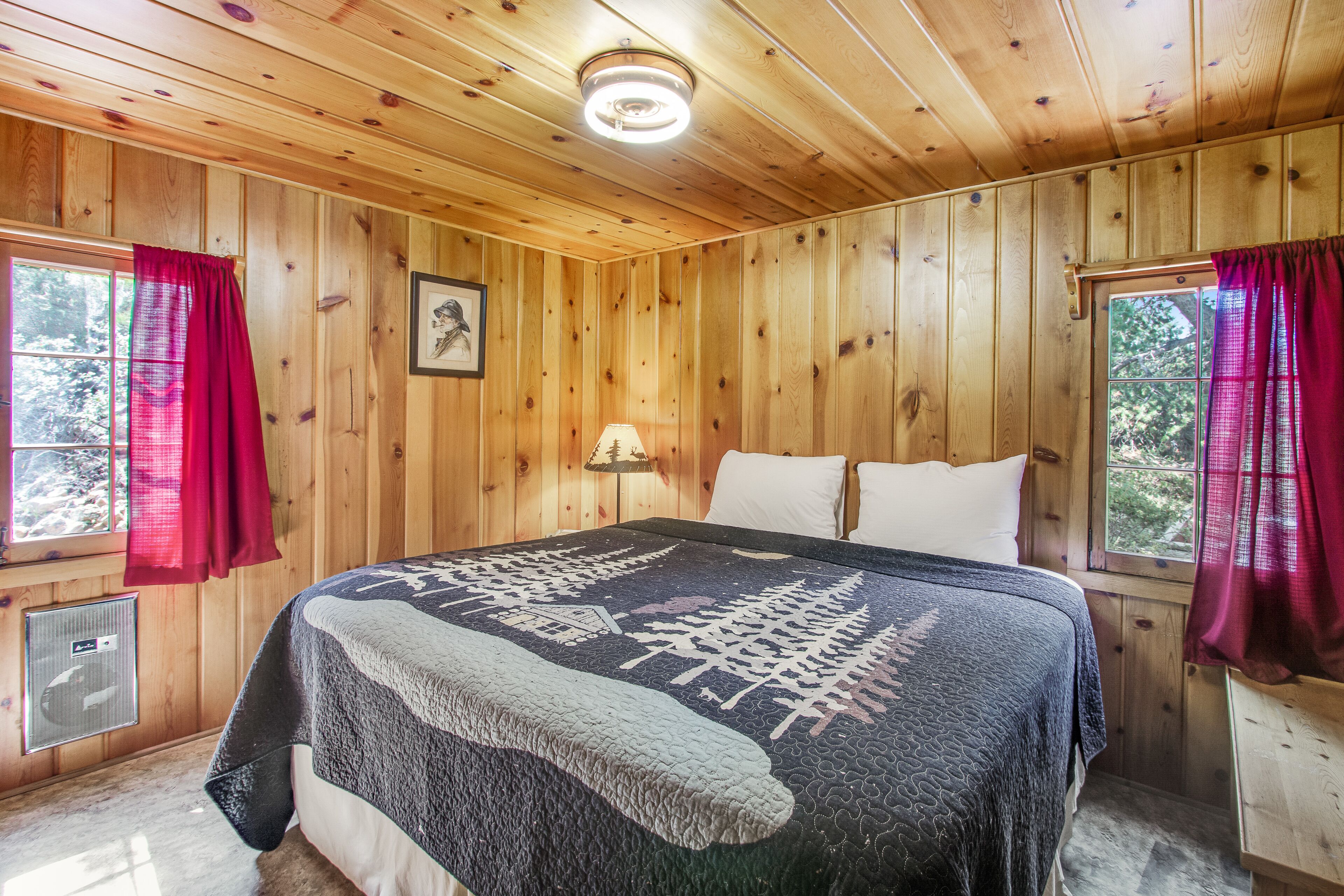 Une chambre dans une cabane en bois, avec un lit, deux fenêtres ornées de rideaux rouges, une lampe fixée au mur et un tableau encadré.