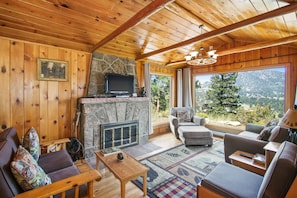 L’intérieur chaleureux d’un chalet avec une cheminée en pierre, des murs en bois et une grande fenêtre donnant sur une vue imprenable sur les montagnes.