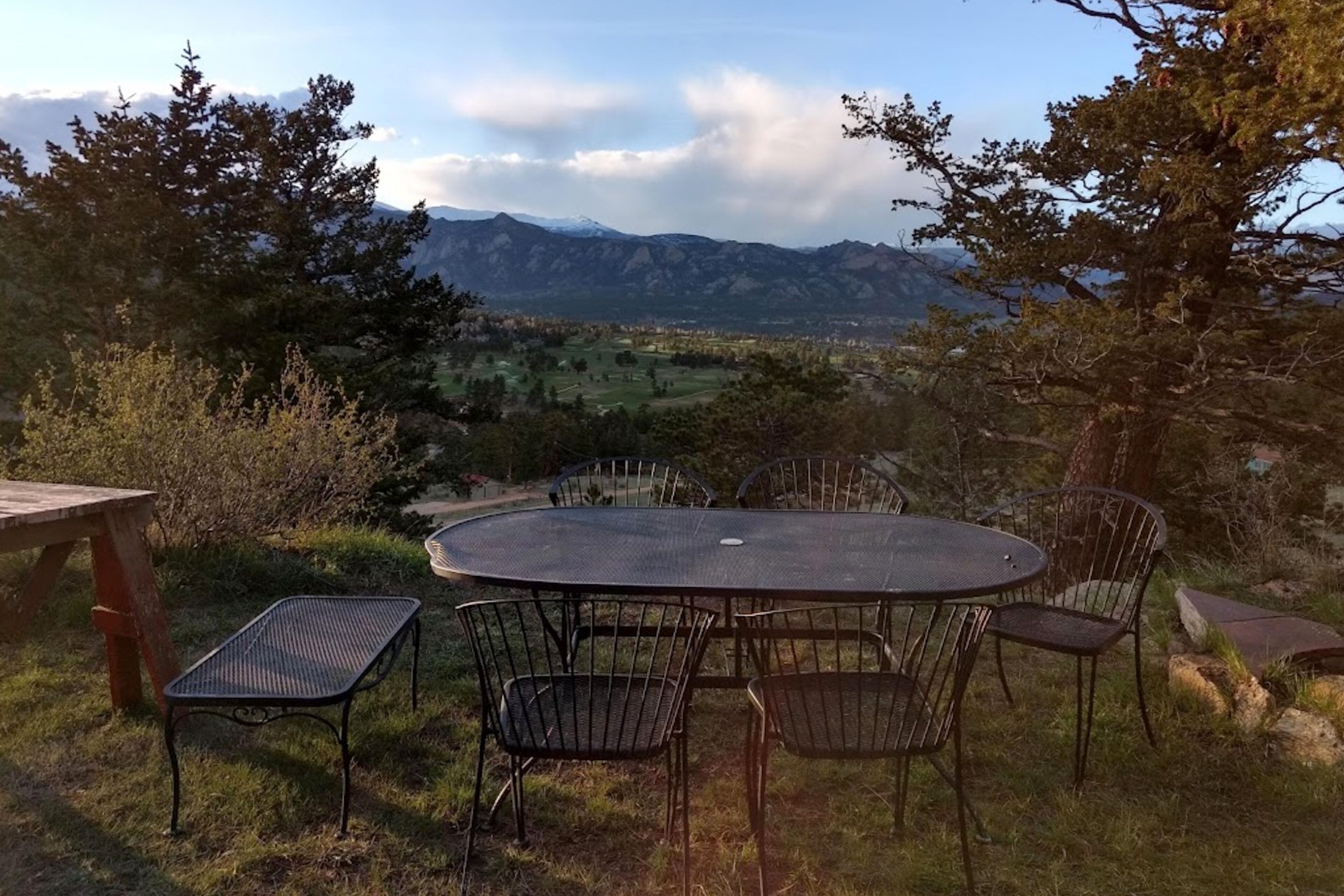 Ein runder Gartentisch mit Metallstühlen, umgeben von Gras und Bäumen, mit Blick auf ein Tal und Berge im Hintergrund.